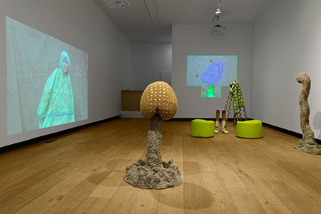 Video projection on left wall of Every worm deserves a mansion video, atomic stink bomb sculpture in foreground, and worm sculpture near 'a wrinkle in the binky' installation in background (smoke, fire and feet with trackpad sculptures. There are also 2 neon green bean bag stumps placed near the trackpad sculpture. 